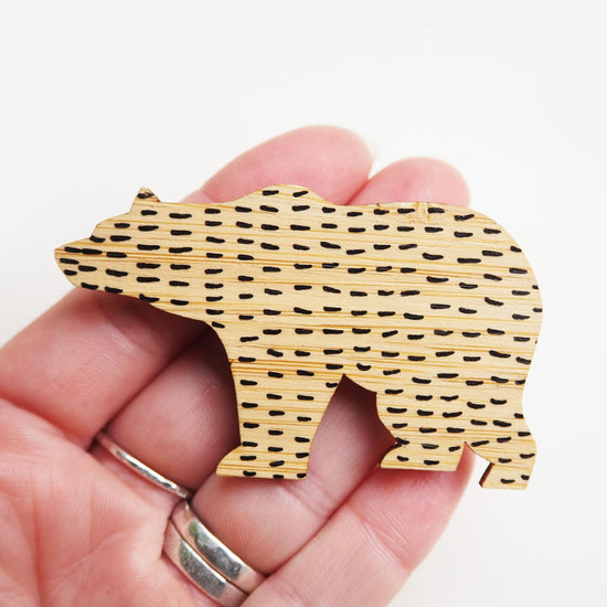 Bamboo bear brooch held at the tip of four fingers. The bear has black short lines in a horizontal pattern all across it.