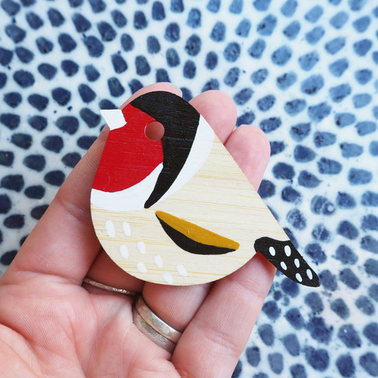 The gold finch brooch resting on the tips of four finger in front of a blue dotted white tile background.