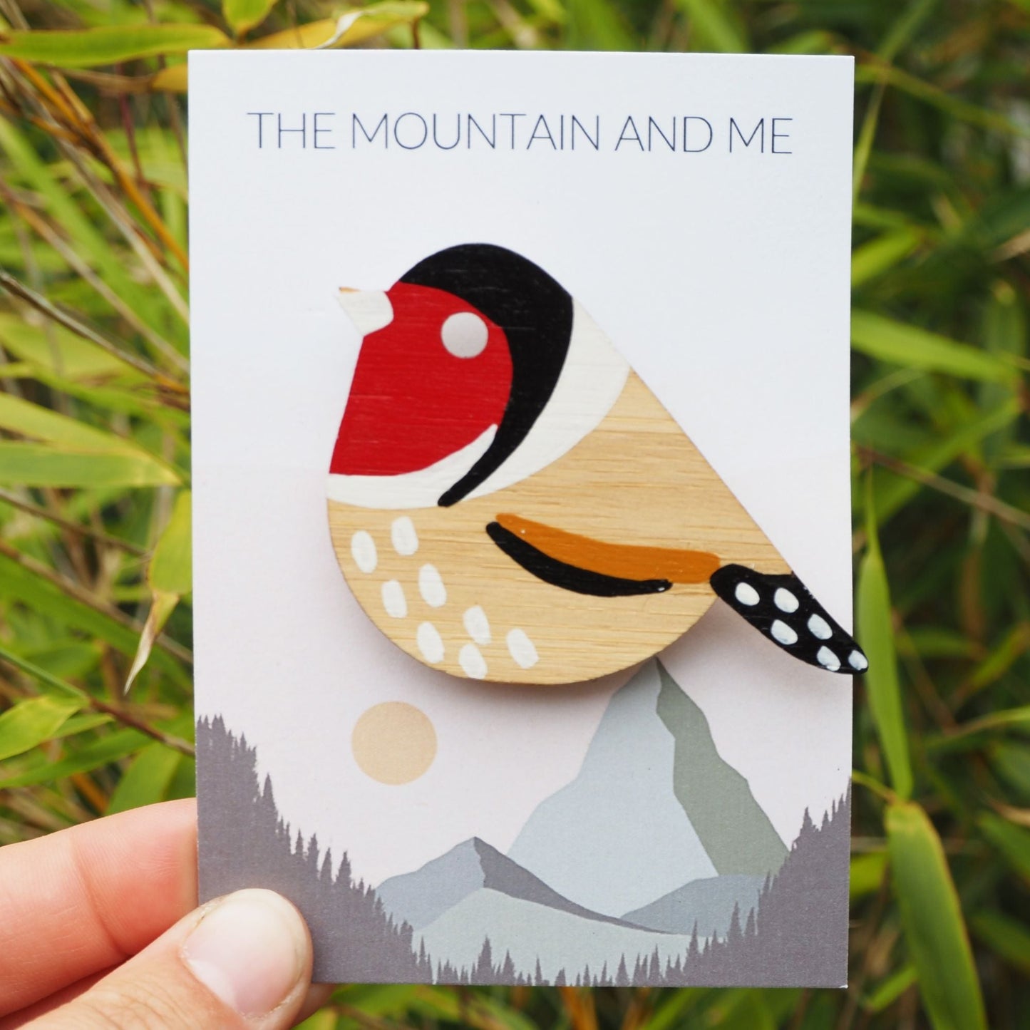 The gold finch brooch on the Mountain and Me branded backing card. It's being held in front of some greenery.