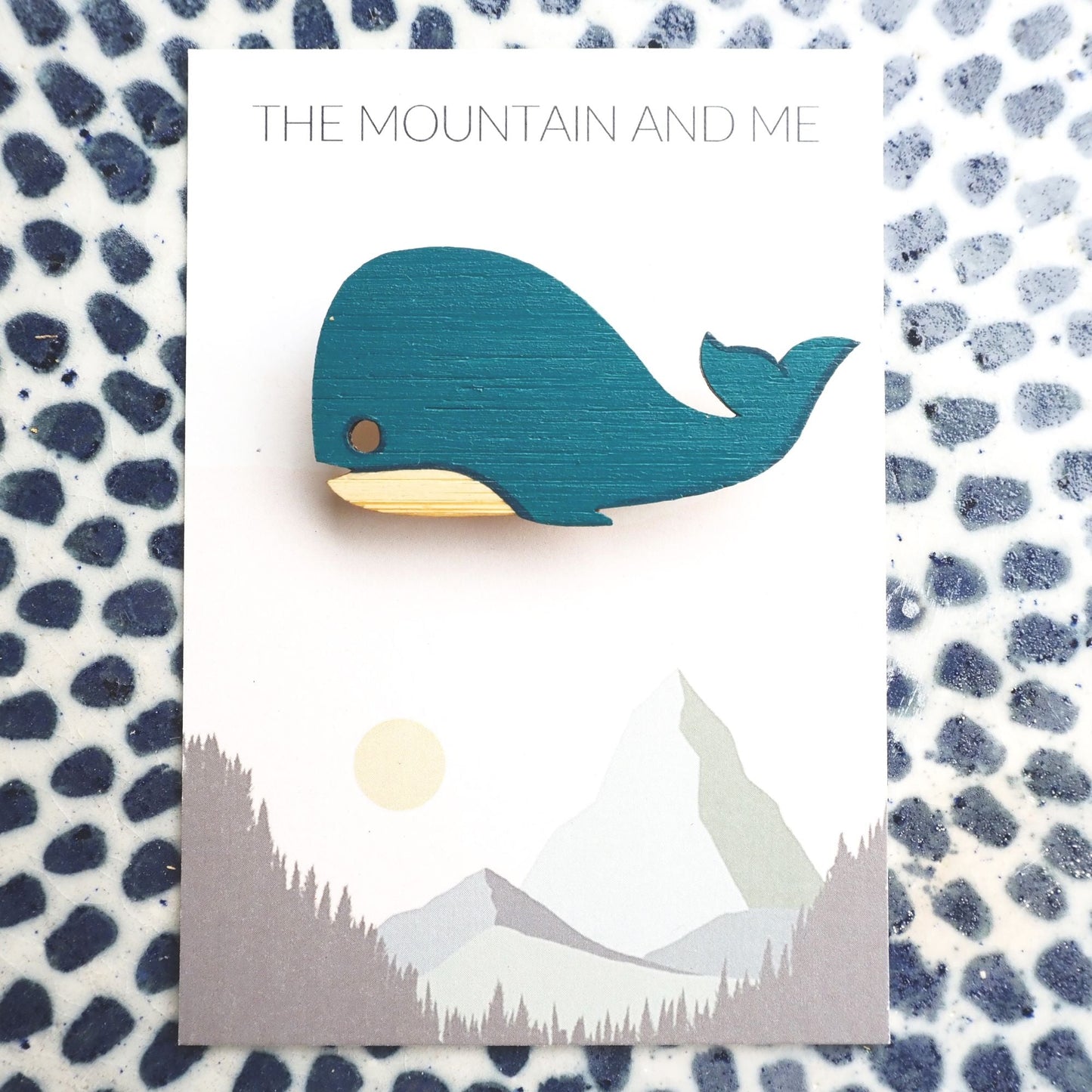 Whale brooch on the Mountain and Me branded backing card. The whale and card is lying on a blue dotted white tile.