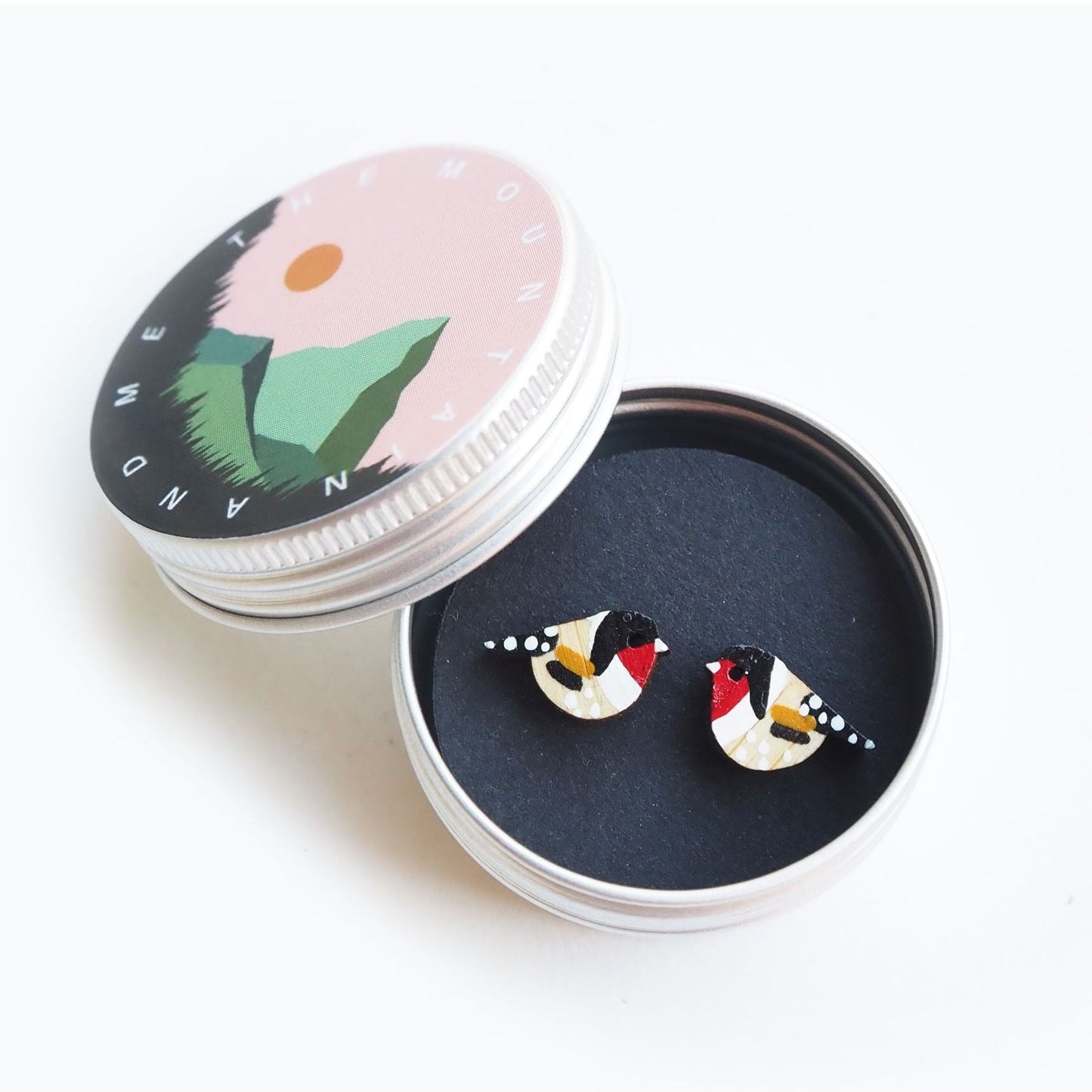 The gold finch earrings inside the screw top tin. The lid is resting against the side of the open tin and has the mountain and me branding on it.