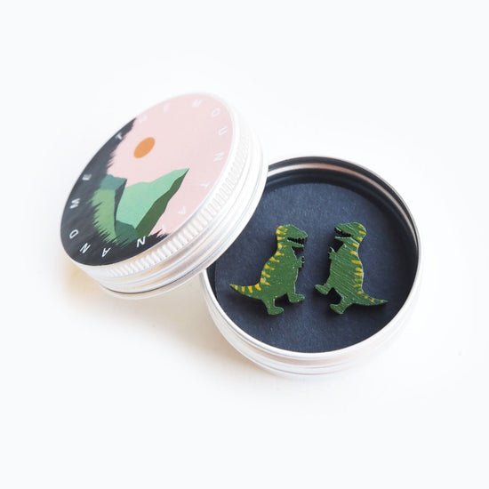 The t rex earrings in the small screw top tin that has mountain and me branding on the lidwhich leans against the side of the open tin.