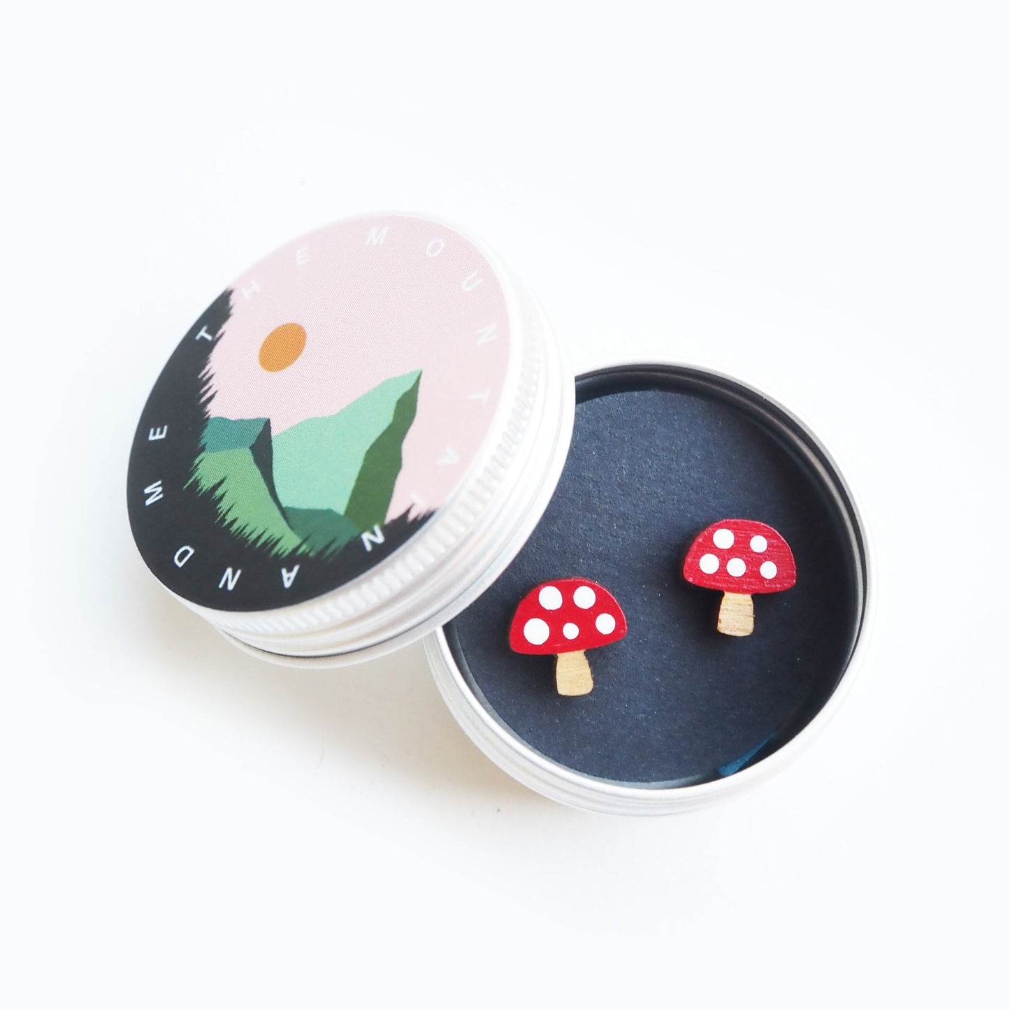The red and white toadstool earrings inside the screw top tin. The lid is resting agaisnt the side of the open tin and has the mountain and me branding on it.