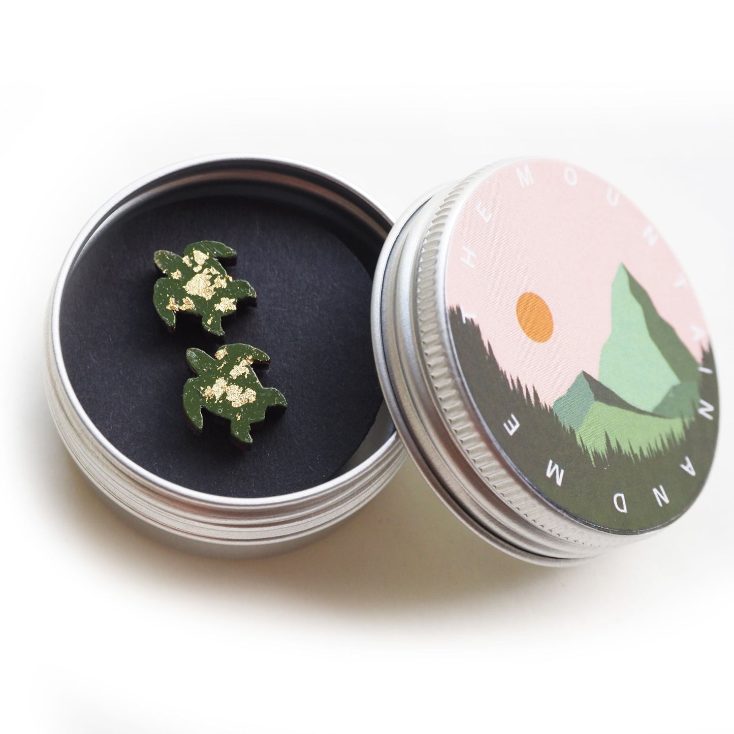 The green and golden turtle earrings inside the screw top tin. The lid is resting against the side of the open tin and has the mountain and me branding on it.