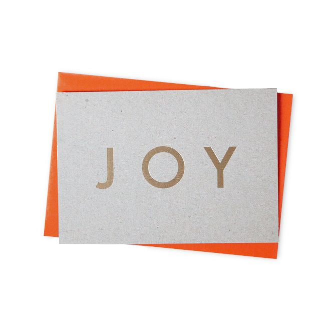Grey card with brass lettering saying, joy. Orange envelope behind the card.