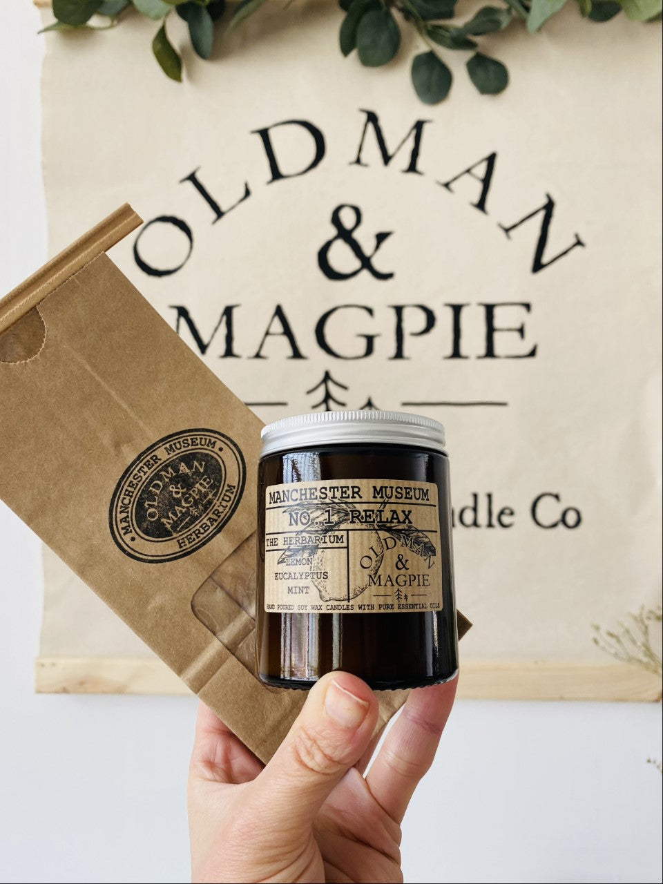 Load image into Gallery viewer, Lifestyle shot with a hand holding the large candle  and the paper bag packaging with an Old Man &amp;amp; Magpie poster in the background.
