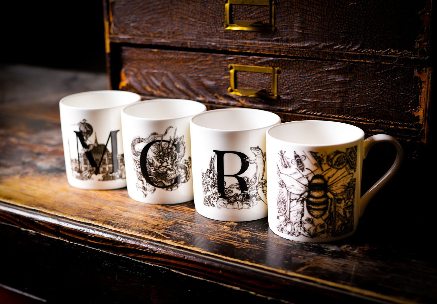 Lifestyle shot of the full mug set from the Sculpts. The four mugs spell out MCR and have the bee mug on the right nearest the viewer. The mugs are standing ona dark brown wooden surface with a brown old file holder behind.