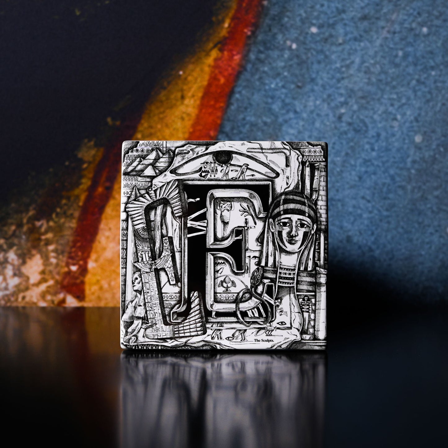 Load image into Gallery viewer, Lifestyle shot of the Egypt and Sudan tile by the Sculpts. The tile is on a reflective black surface and standing in front of a banner image from which only some dark blue and golden and red lines can be seen.
